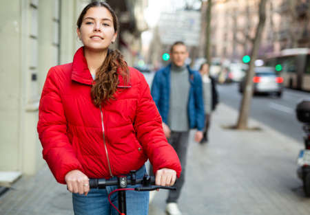 Attractive girl in jacket standing with electric scooter outdoors, ready to ride on city streetsの写真素材