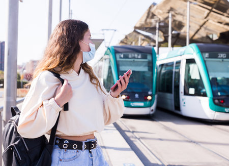 Woman in mask holding smartphone while waiting for tramの写真素材
