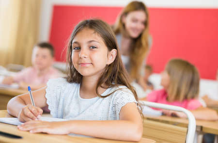 Smiling tween schoolgirl writing in workbook at lesson in classの写真素材