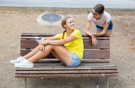 Two girls friends sitting on bench in summer park chatting happily having fun