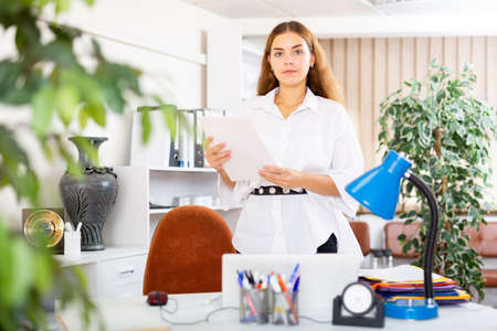Confident female office employee standing with papers near workplaceの写真素材