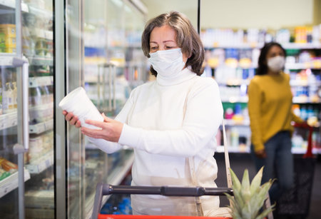 Woman in protective mask with shopping cart choosing groceriesの写真素材