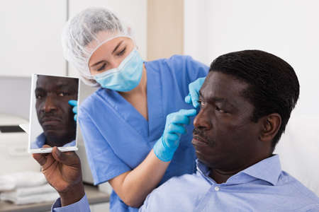 Woman beautician in mask examining face skin of afro american male patientの写真素材