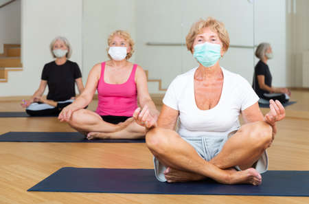 Aged woman in protective mask sitting in lotus positions during group yoga trainingの写真素材