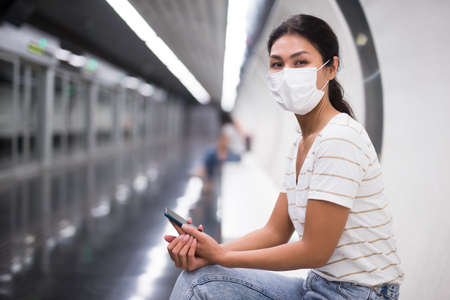 Woman in face mask with smartphone sitting on bench in subway stationの写真素材