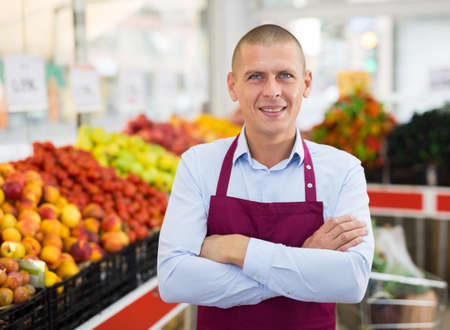 Confident greengrocery owner near shelves with fruits and vegetablesの写真素材