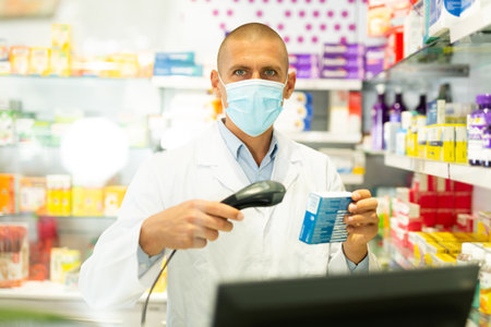 Portrait of pharmacist in medical mask working at the cash register in pharmacy - scans barcode on medicine packageの写真素材