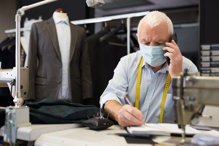Mature tailor wearing protective mask talking on a mobile phone and taking notes in notebook