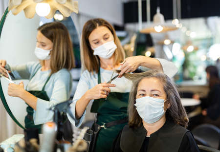 Woman hairdresser in protective mask cutting hair of elderly female clientの写真素材