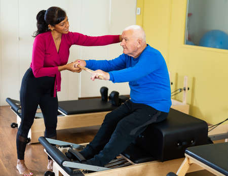 Senior man exercising with stick on pilates reformer under supervision of Hispanic female instructorの写真素材