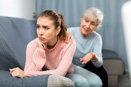 Senior woman calming chagrined adult daughter in home living roomの写真素材