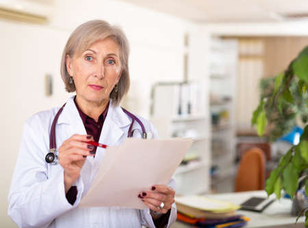 Elderly female doctor with papers studying clinical diagnosis of patientの写真素材