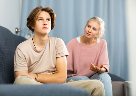 Upset and offended teenager sitting on sofa while mother soothing himの写真素材