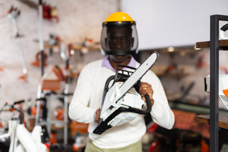 African-american man in protective helmet and with chainsaw in hands in gardening tools shop.