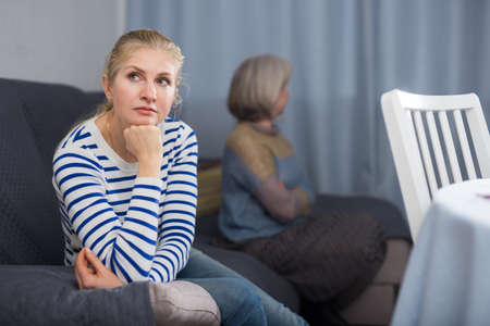 Elderly mother and adult daughter sit on the couch turned away from each other after domestic quarrelの写真素材
