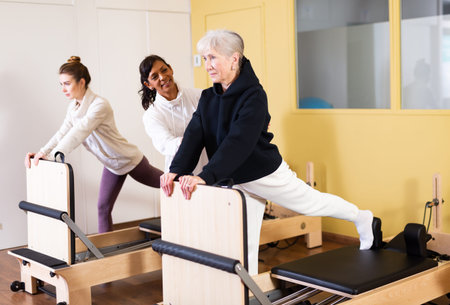 Elderly woman practicing pilates on reformer with Hispanic female trainer