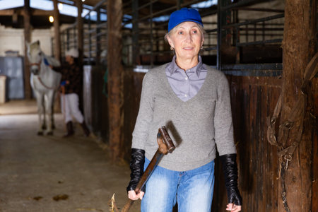 Smiling elderly woman carrying pitchfork to stack hay for horses in stableの写真素材