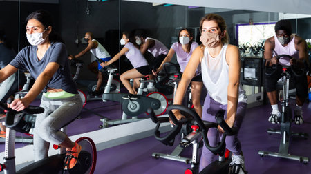 Men and women wearing protective masks ride stationary bike in fitness club