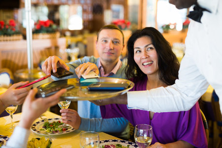 African american waiter collecting phones of cheerful company in restaurant