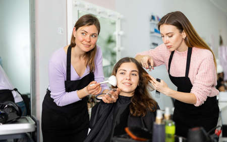 Professional female stylists making hairstyle and makeup to young womanの写真素材