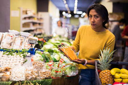 Interested hispanic woman choosing fresh carrots in supermarketの写真素材