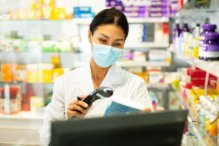 Portrait of female pharmacist in medical mask working at the cash register in pharmacy - scans barcode on medicine packageの写真素材