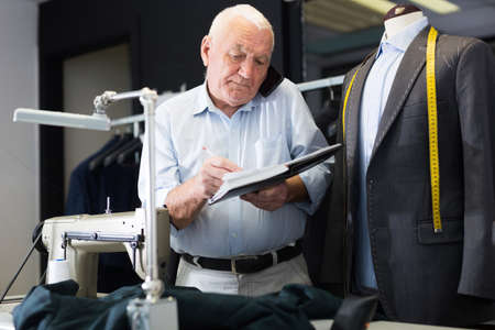 Professional tailor man talking on mobile phone and making notes in notebookの写真素材