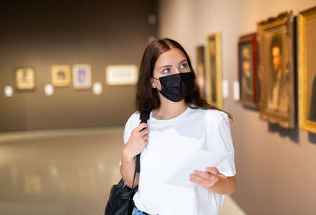 Focused girl in a protective mask looks at the paintings in the museum hallの写真素材
