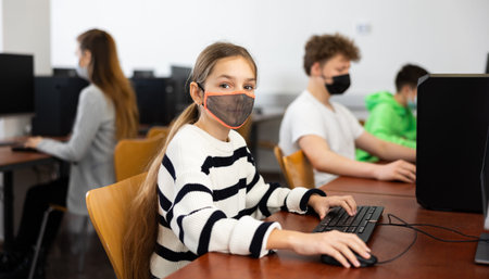 Group of young girls and boys in face masks sitting in computer classroom of library
