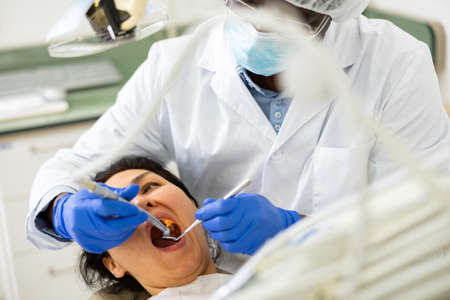 Dentist in face mask inspects female patient teeth with mirrorの写真素材