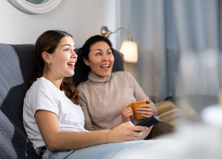 Two female friends watching TV while sitting on sofa at homeの写真素材