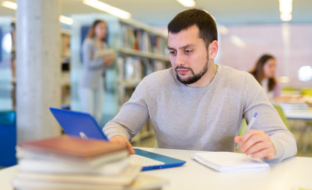 Middle aged man reading books and working on laptop in library