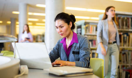 Focused woman working on laptop at libraryの写真素材