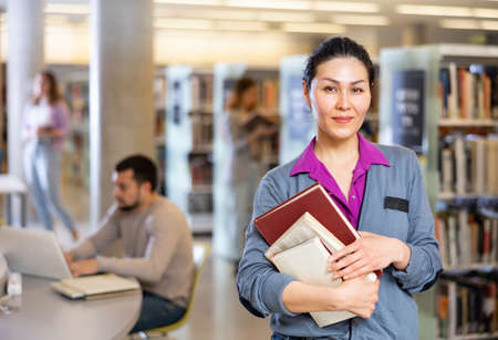 Woman reading book while standing near bookshelves in libraryの写真素材