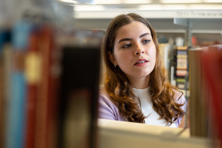 Student girl in the library is looking for the right book on the shelf of the rack