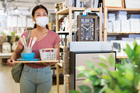 Female customer in protective mask holding purchases and walking in household goods store