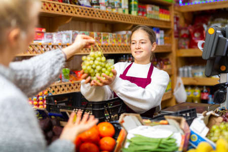 Girl who works part-time as a trainee saleswoman handing grape to customer after payment at checkoutの写真素材