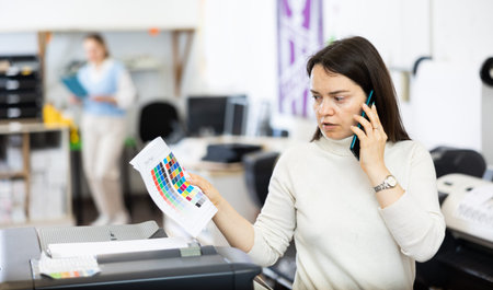 Woman employee of printing shop holds a color chart in her hands and consults on mobile phone