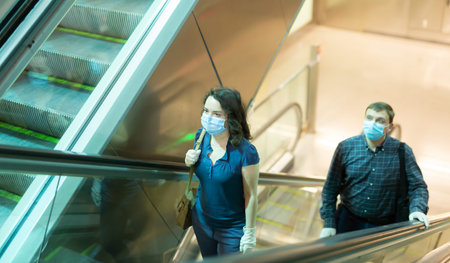 Focused people in medical masks and rubber gloves lifting on escalator keeping distance to prevent...