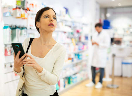 Woman chooses medical products in pharmacy, checking the list on smartphoneの写真素材
