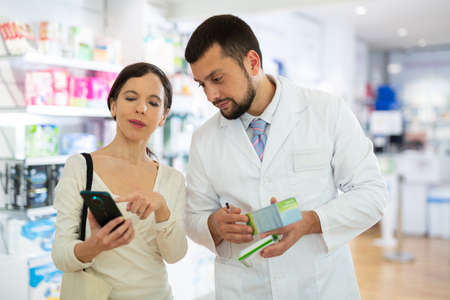 Female shopper shows name of drug on smartphone screen. Pharmacist helps to find medicine in pharmacyの写真素材
