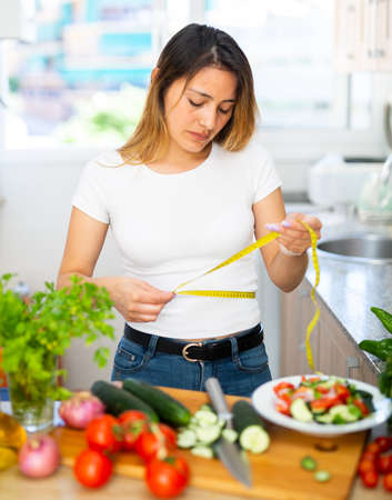 Upset housewife measures waist before eating vegetable saladの写真素材