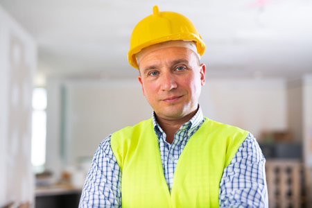 Portrait of smiling worker in yellow vest and hard hat inside cottage under renovation