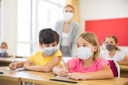 Preteen pupils in protective masks studying in classroom with female teacher