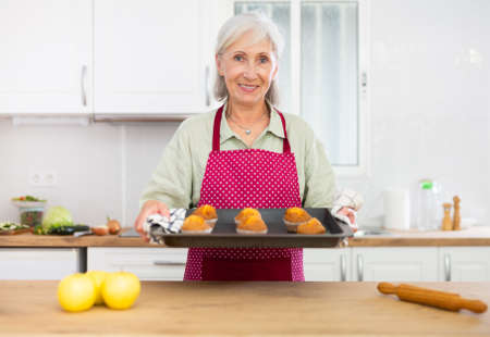 Smiling senior woman in apron holding sheet pan cupcakesの写真素材
