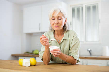 Mature woman eating tasty yogurt in kitchenの写真素材