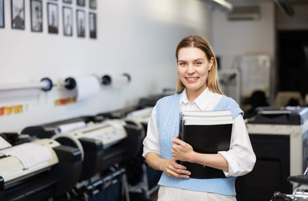 Portrait of positive female employee of a printing house with notebooks and projects in hands