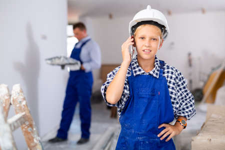 Little builder boy in blue overalls talking on a mobile phone in room being renovatedの写真素材