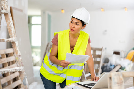 Woman builder checking project documentation and using laptop