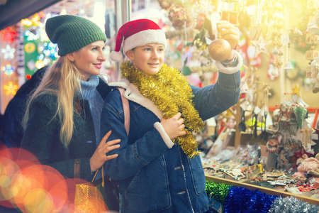 Happy teenage girl with mother choosing Christmas decoration at street fairの写真素材